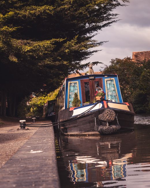 A narrowboat moored along the Hertford Union Canal, featuring a vibrant blue and red exterior with decorative stained glass windows. The boat's front is secured with thick black mooring ropes attached