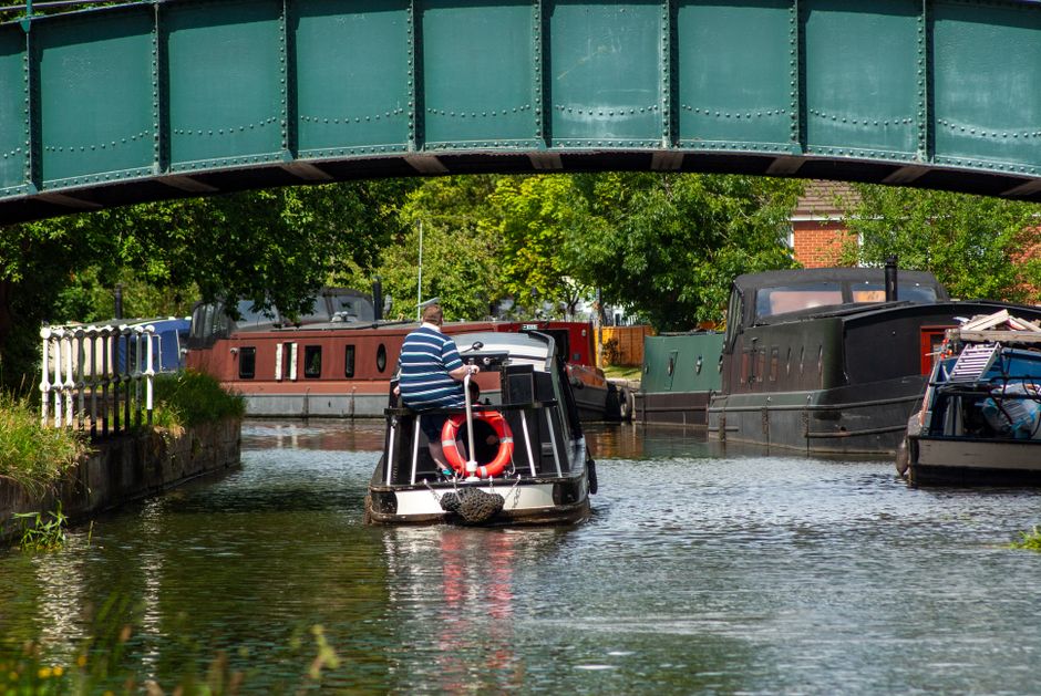 A narrowboat moored along the Hertford Union Canal, featuring a vibrant blue and red exterior with decorative stained glass windows. The boat's front is secured with thick black mooring ropes attached